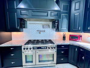 Gourmet kitchen featuring a professional-grade stainless steel range with double ovens, dark cabinetry with decorative hardware, light granite countertops, a subway tile backsplash, and a pot filler