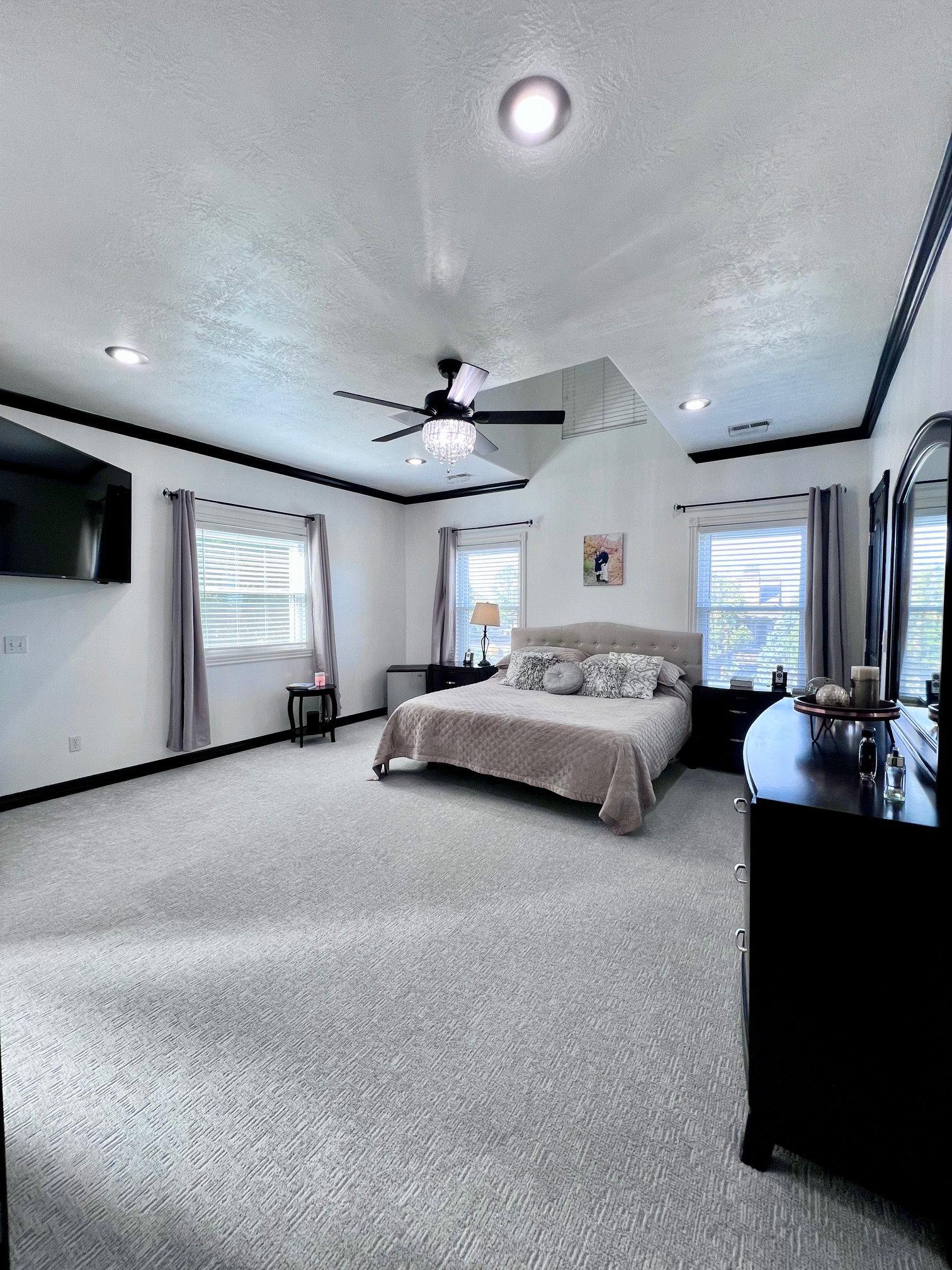 Spacious bedroom featuring light grey patterned carpeting, multiple windows with blinds, a ceiling fan with integrated lighting, recessed lighting, and contrasting dark crown molding