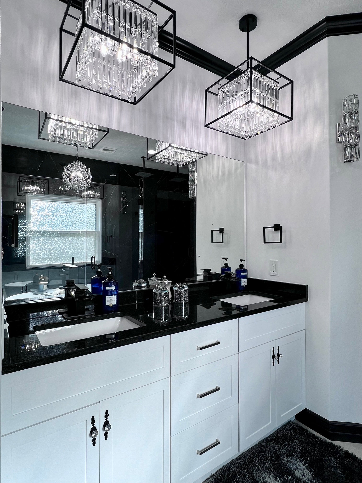 Dual crystal chandeliers illuminate a vanity featuring a black countertop, two undermount sinks, and white cabinetry with varied hardware