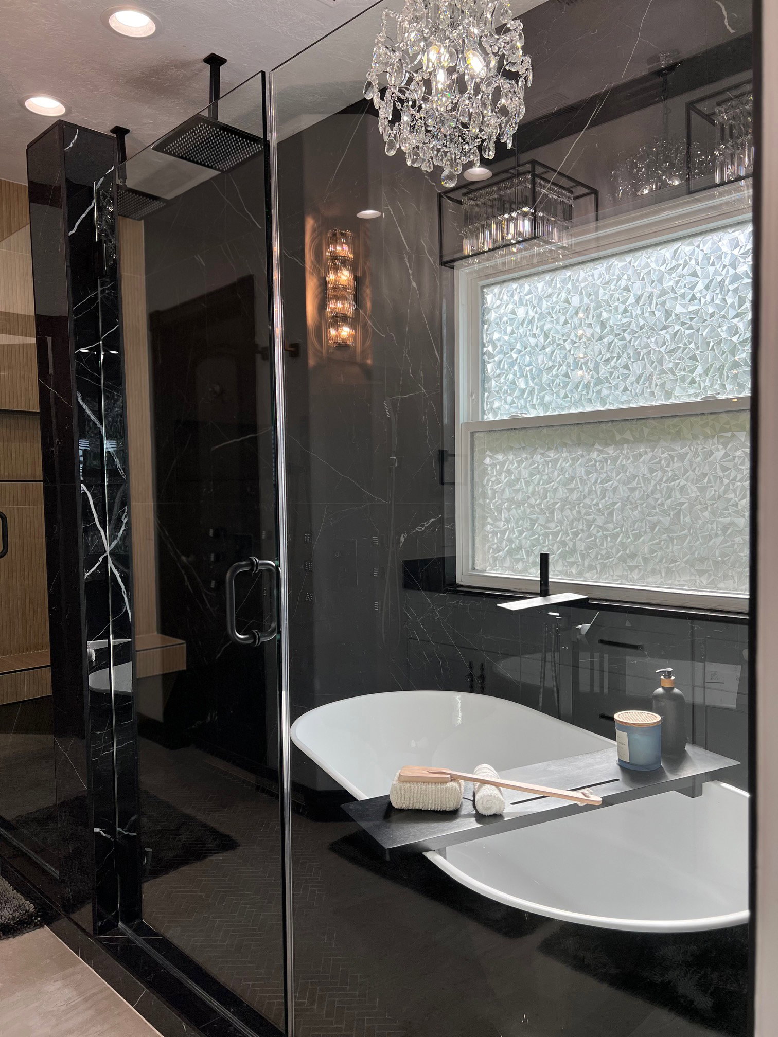 Contemporary bathroom featuring a freestanding soaking tub with a floor-mounted faucet, a spacious glass-enclosed shower with a rainfall showerhead, black marble-look wall tiling, and a decorative crystal chandelier