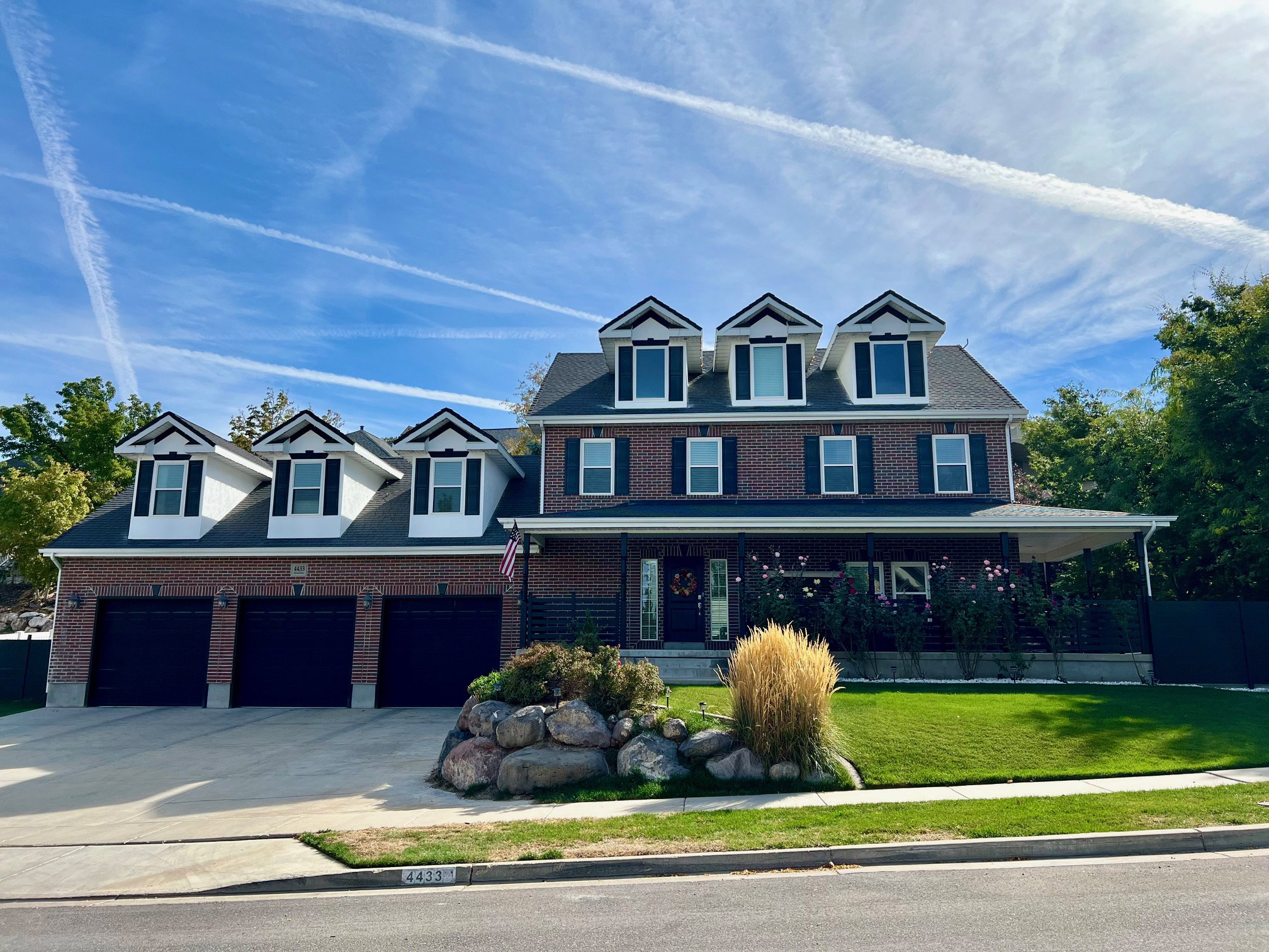 Brick facade residence featuring multiple dormer windows, a covered front porch, an attached three-car garage, manicured front lawn, and mature landscaping