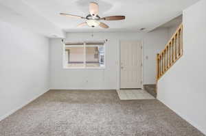Carpeted living space featuring a ceiling fan with integrated lighting, a multi-pane window, a paneled entry door, and a staircase with wood-finish railing
