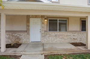 Covered front porch featuring a white entry door with brass hardware, brick and stucco exterior, a large window with white framing, an exterior wall light fixture, and concrete flooring