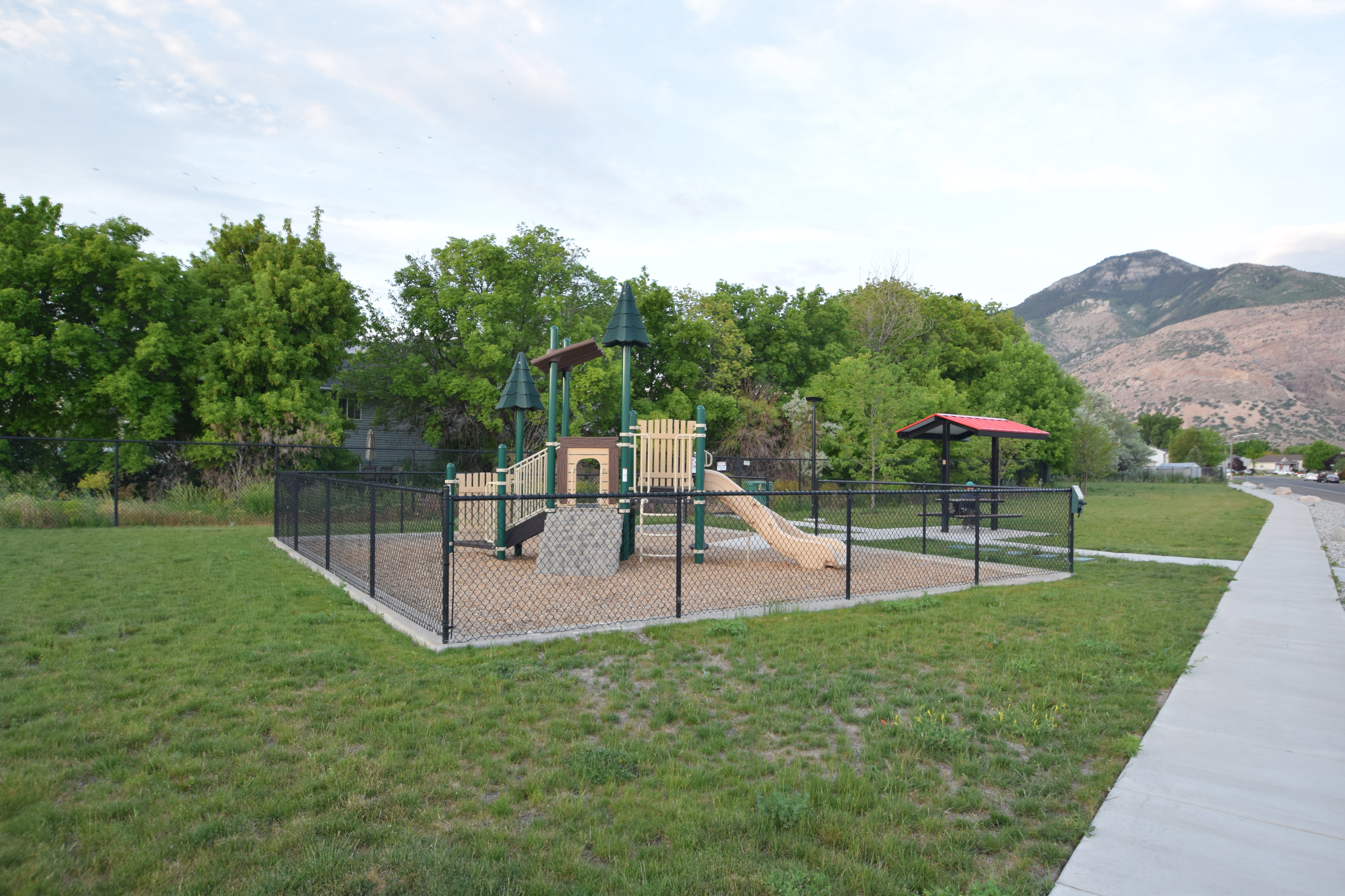 Fenced playground area featuring a play structure with slide, a separate covered picnic area, and surrounding green space
