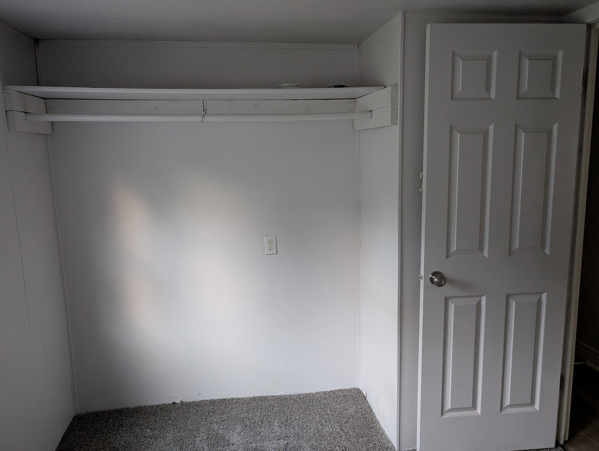Built-in closet with a hanging rod and shelf above, set on carpeted flooring, adjacent to a six-panel interior door with a silver-toned knob