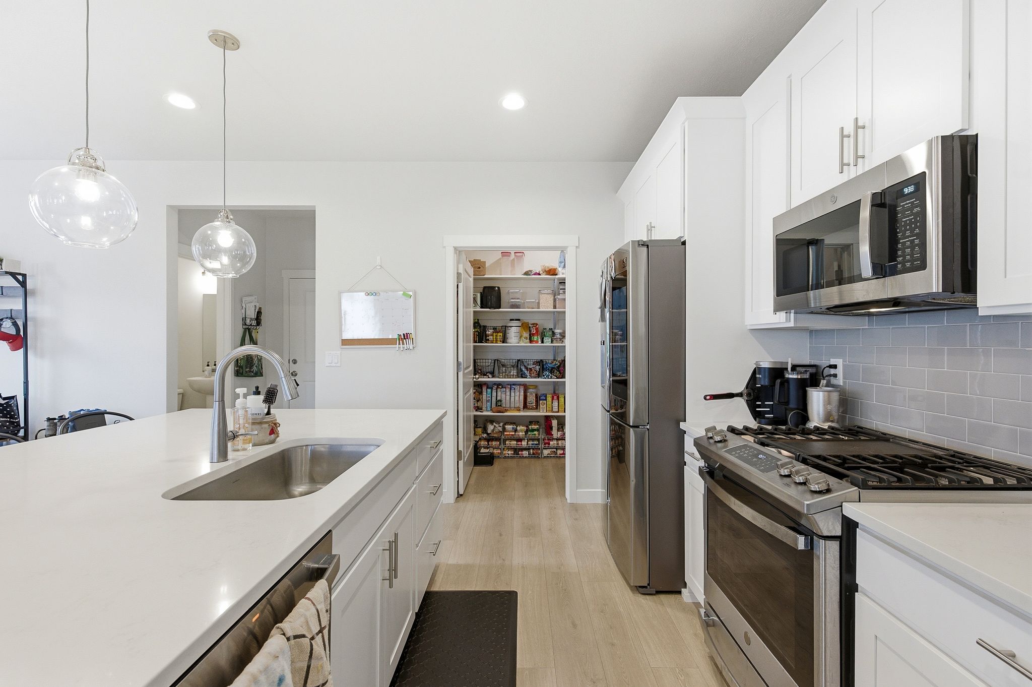 Bright kitchen featuring a large island with an undermount sink, quartz countertops, white cabinetry with contemporary hardware, stainless steel appliances, and a spacious pantry with shelving