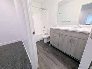 Bathroom featuring wood-finish flooring, a white vanity with a single basin sink, a large mirror, a white toilet, and a white shower-tub combination with chrome fixtures