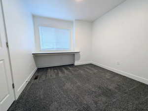 Room featuring a built-in floating desk, a window with horizontal blinds, neutral white walls, and dark gray carpeting