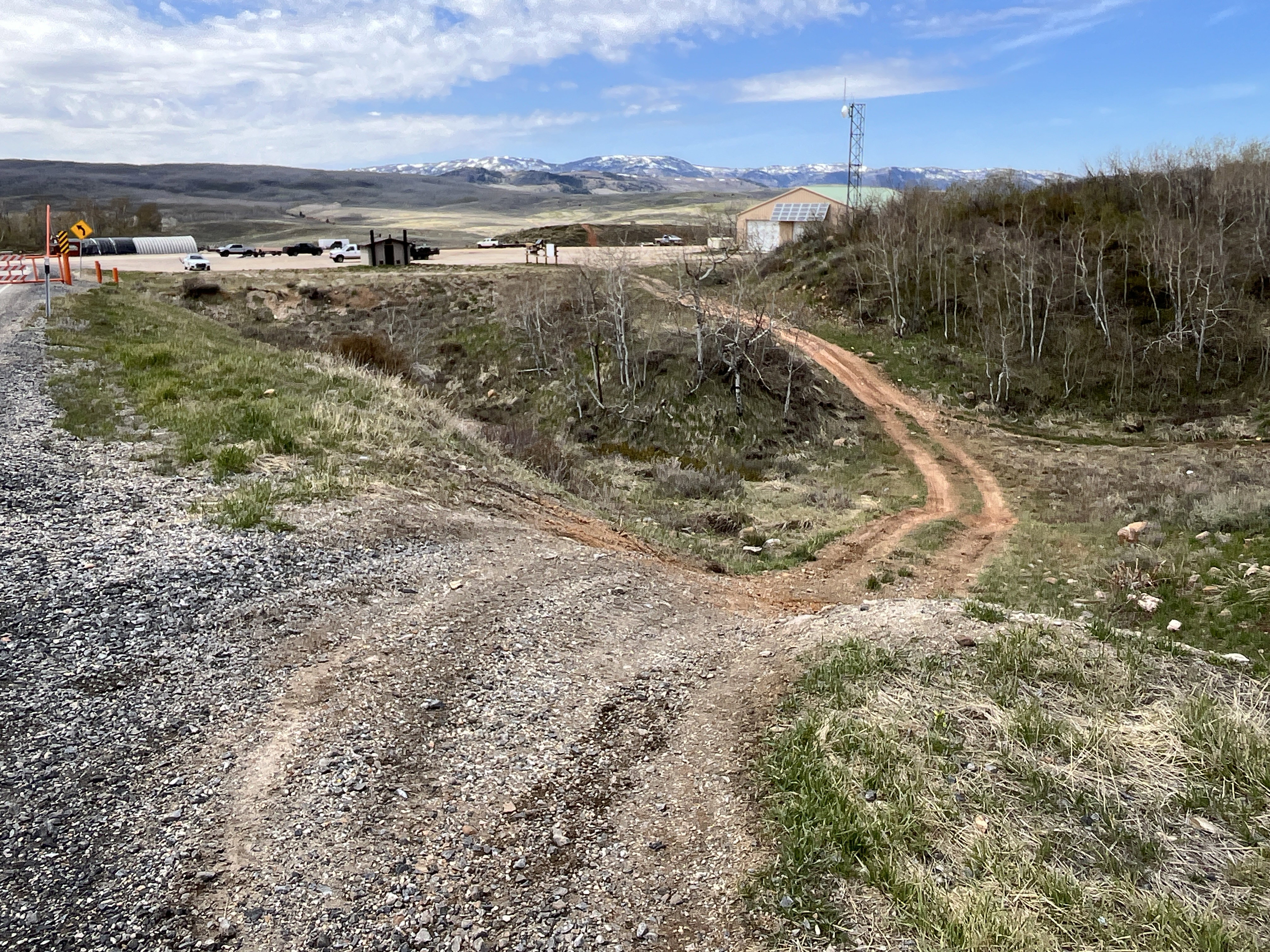Gravel road with a dirt path descending into a natural landscape featuring sparse trees, distant snow-capped mountains, and an industrial-style building