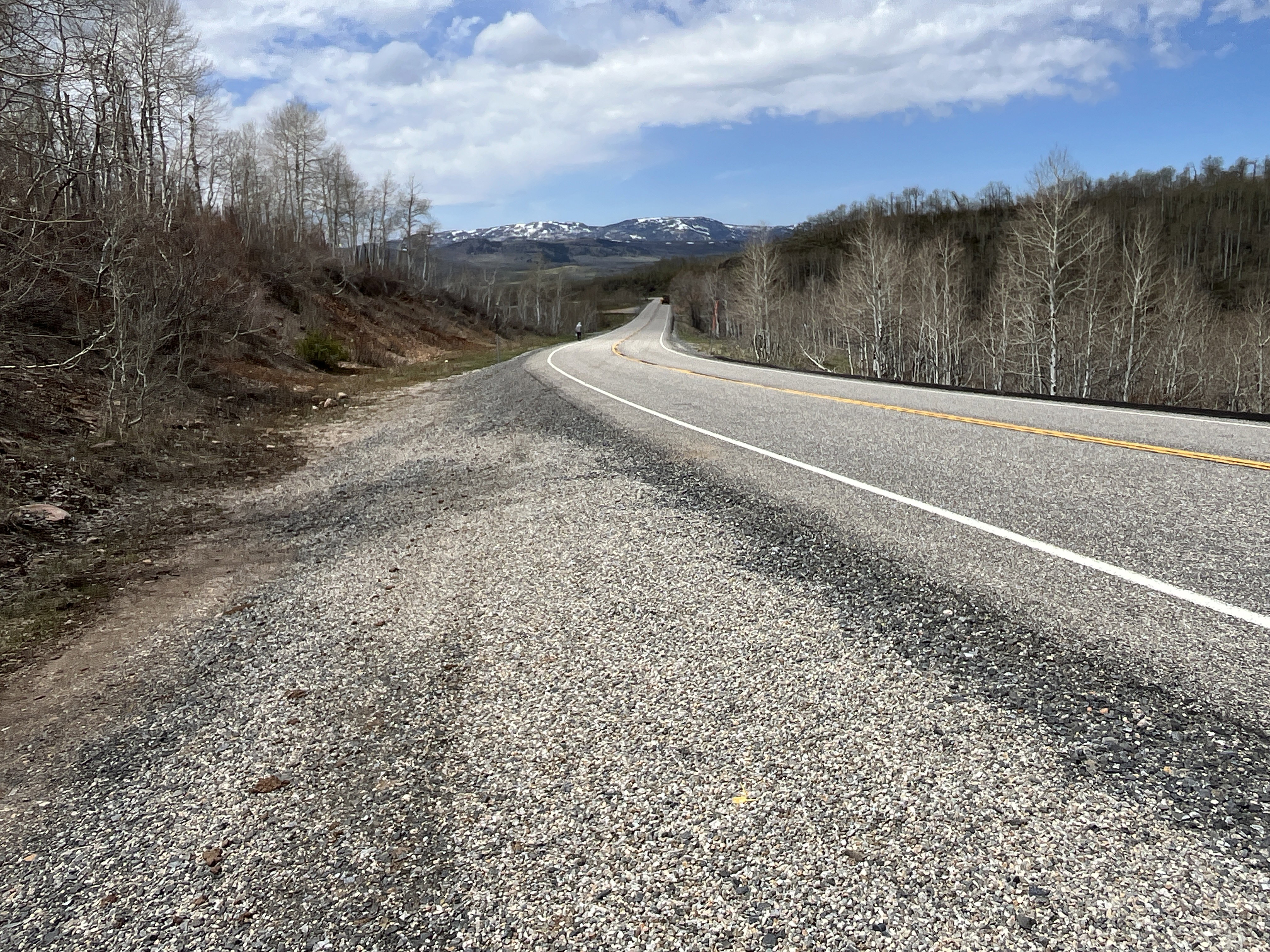 Paved roadway with a gravel shoulder, flanked by wooded hillsides featuring deciduous trees