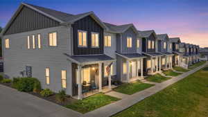 Modern townhome featuring a covered front porch, vertical board and batten siding, horizontal siding, a gray shingle roof, and a concrete walkway bordered by green lawn
