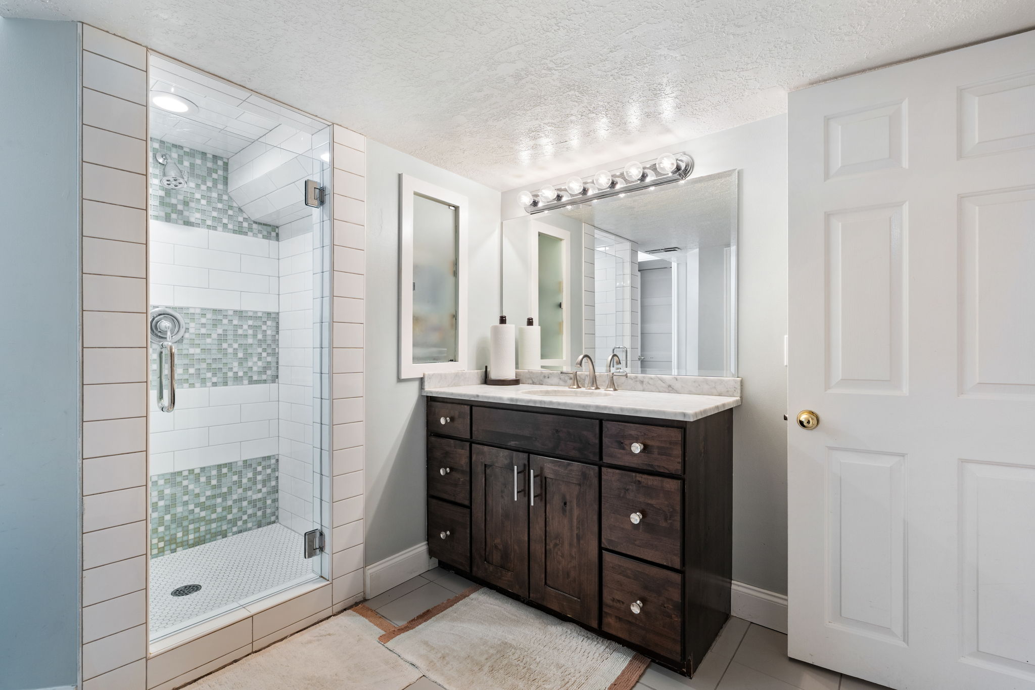 Bathroom featuring a glass-enclosed shower with subway tile and mosaic accents, a dark wood-finish vanity with a light-toned countertop, a large mirror, and a wall-mounted light fixture