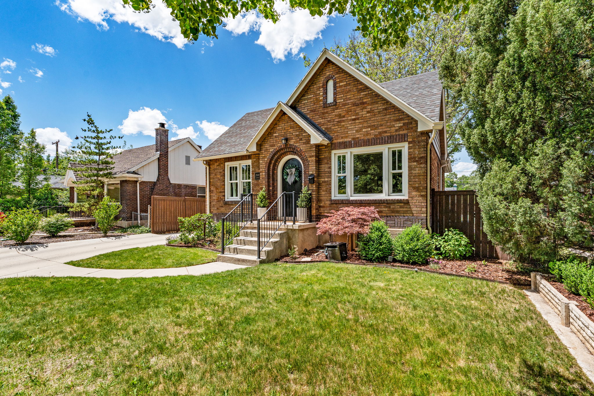 Brick exterior with gabled roofline, arched front entry, concrete steps with wrought iron railings, mature landscaping, and an established lawn