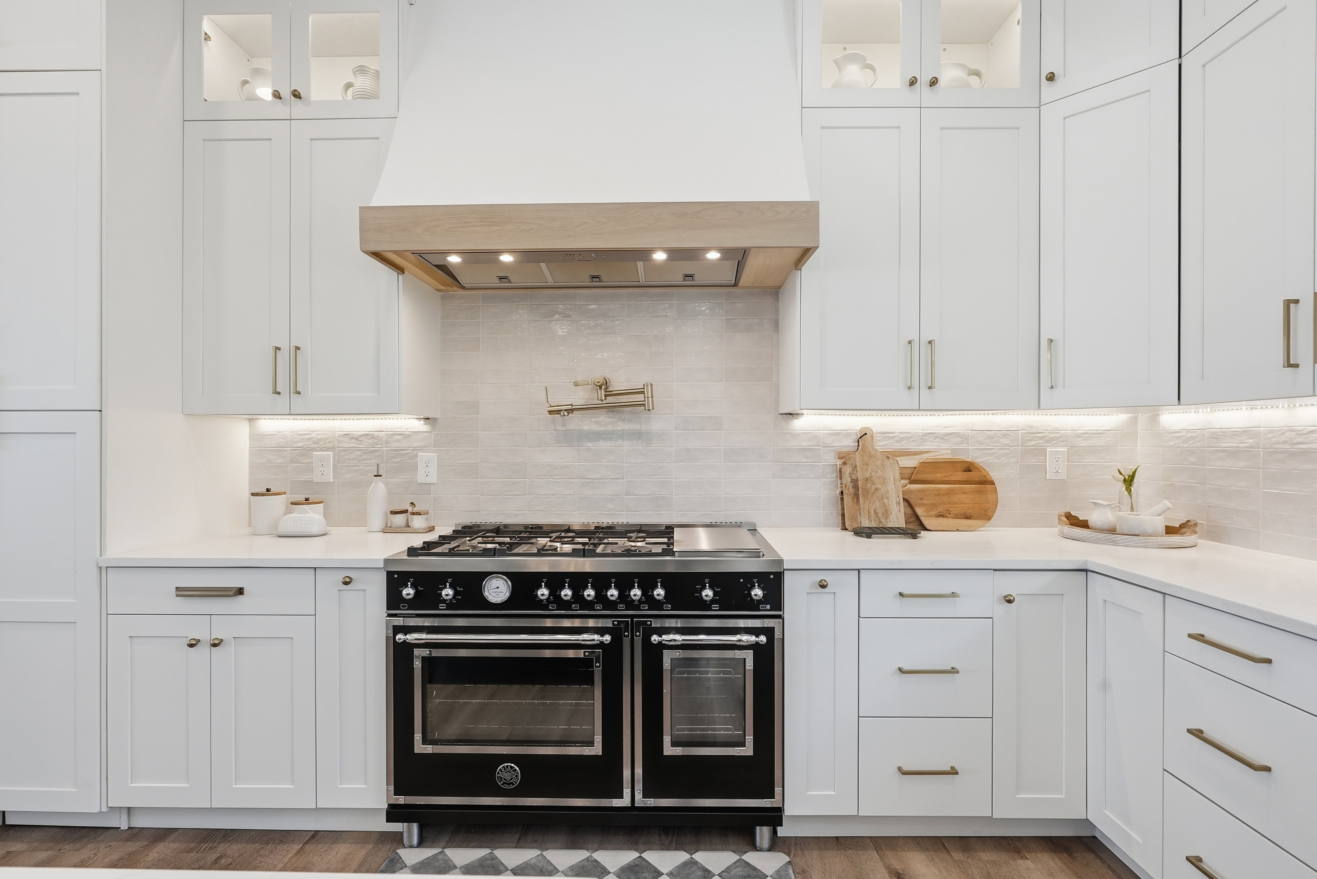 Gourmet kitchen featuring a professional-grade black range, custom wood-paneled range hood, white shaker cabinetry with gold-tone hardware, textured tile backsplash, and under-cabinet lighting