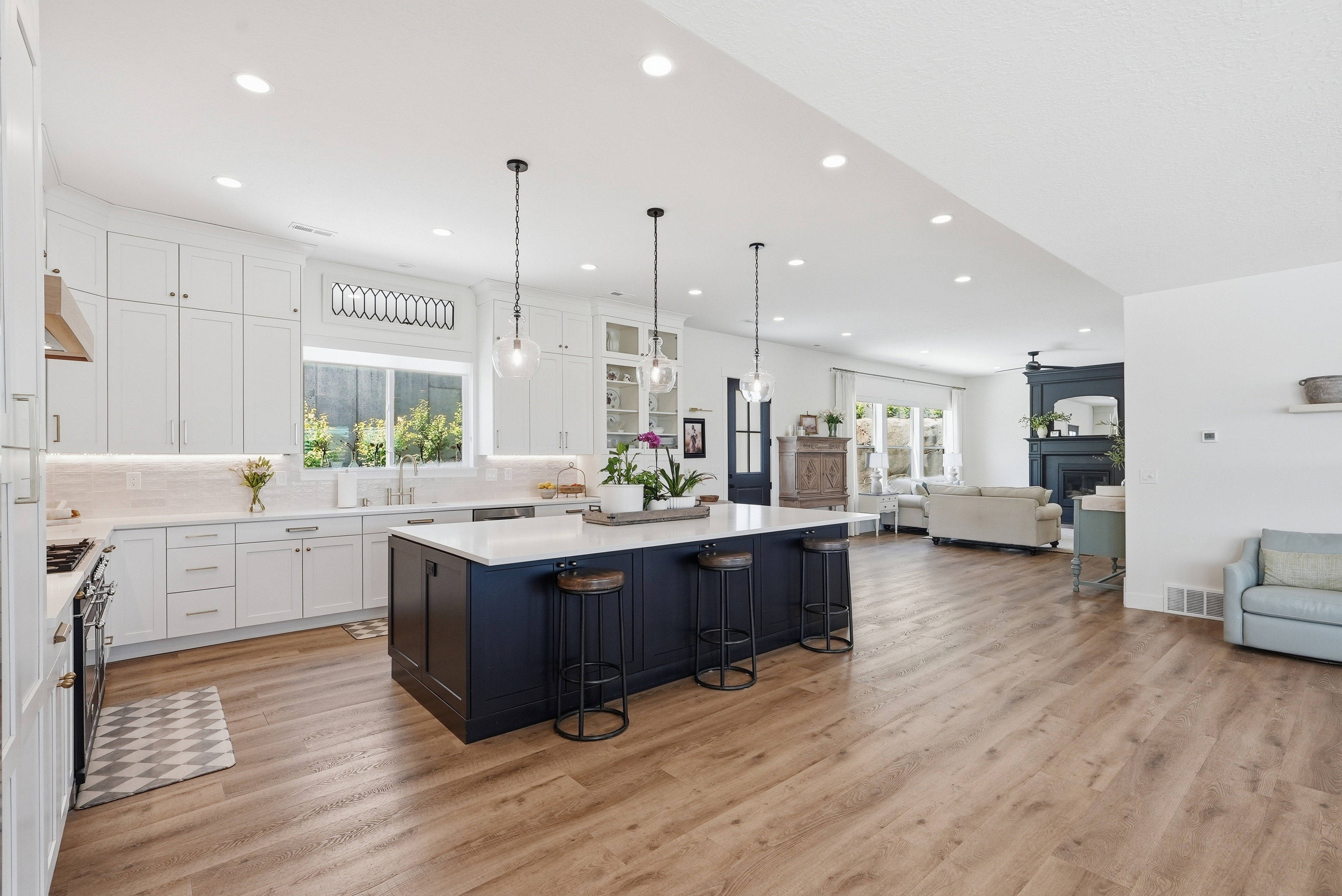 Spacious kitchen featuring an expansive island with seating, white shaker cabinetry, quartz countertops, and wood-finish flooring extending into the living area with a dark-toned fireplace surround