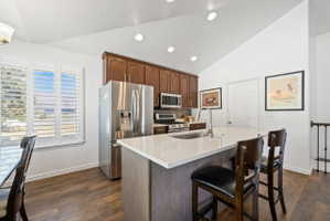 Kitchen featuring stainless steel appliances, light stone counters, a kitchen breakfast bar, a center island with sink, and dark wood-style flooring