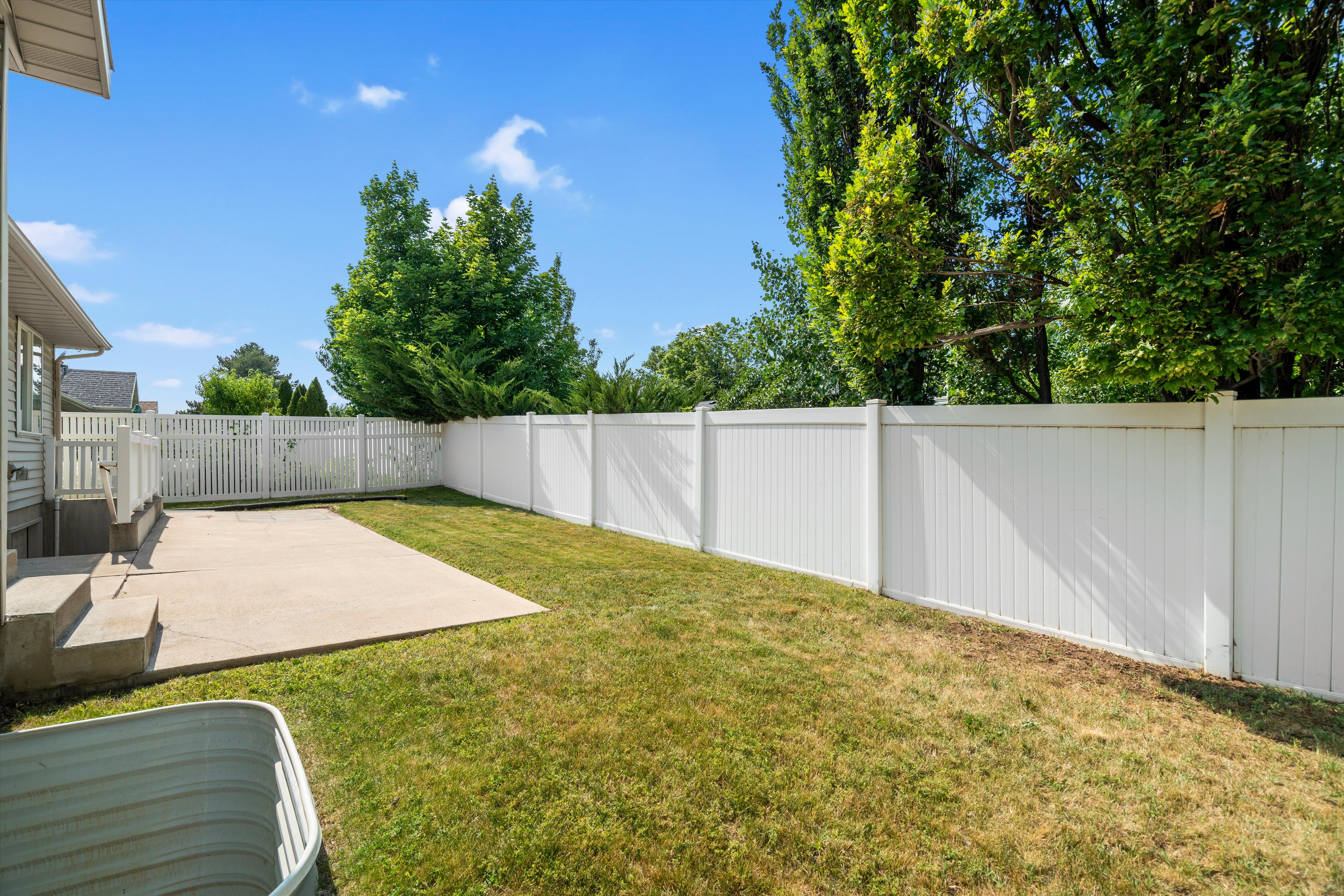 Expansive backyard featuring a concrete patio, a full white vinyl privacy fence, and mature trees surrounding the perimeter