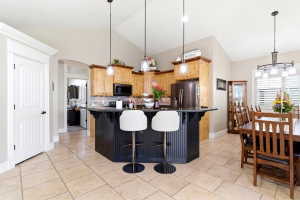 Open-concept kitchen and dining area featuring a dark-toned island with beadboard paneling, light wood cabinetry, stainless steel appliances, a tile backsplash, and light-toned tile flooring
