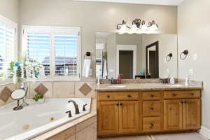 Bathroom featuring a corner soaking tub with tile surround, wood-finish double vanity with stone countertop, and plantation shutters