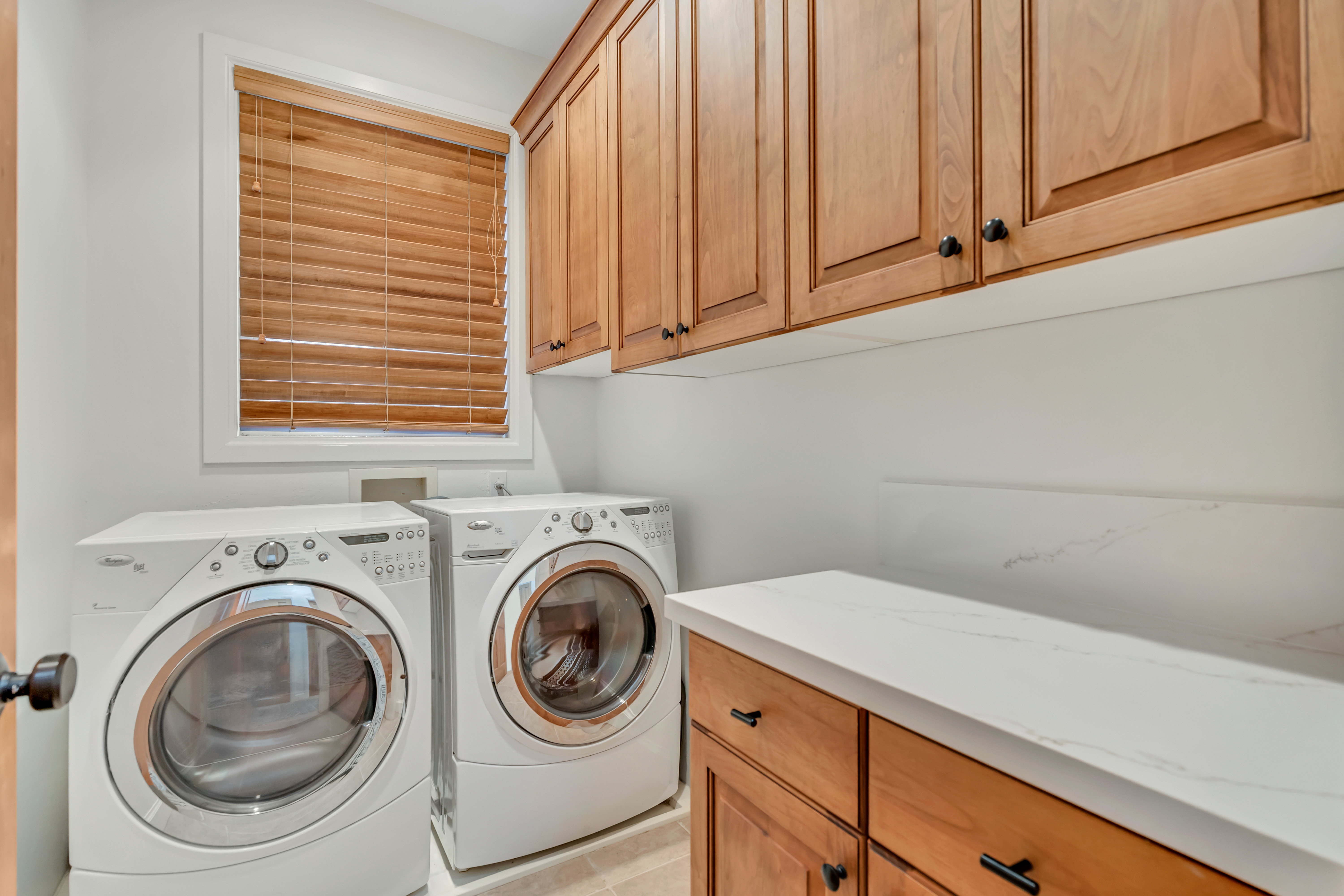 Dedicated laundry room featuring light wood cabinetry, white countertops, a window with wood blinds, and light-colored flooring