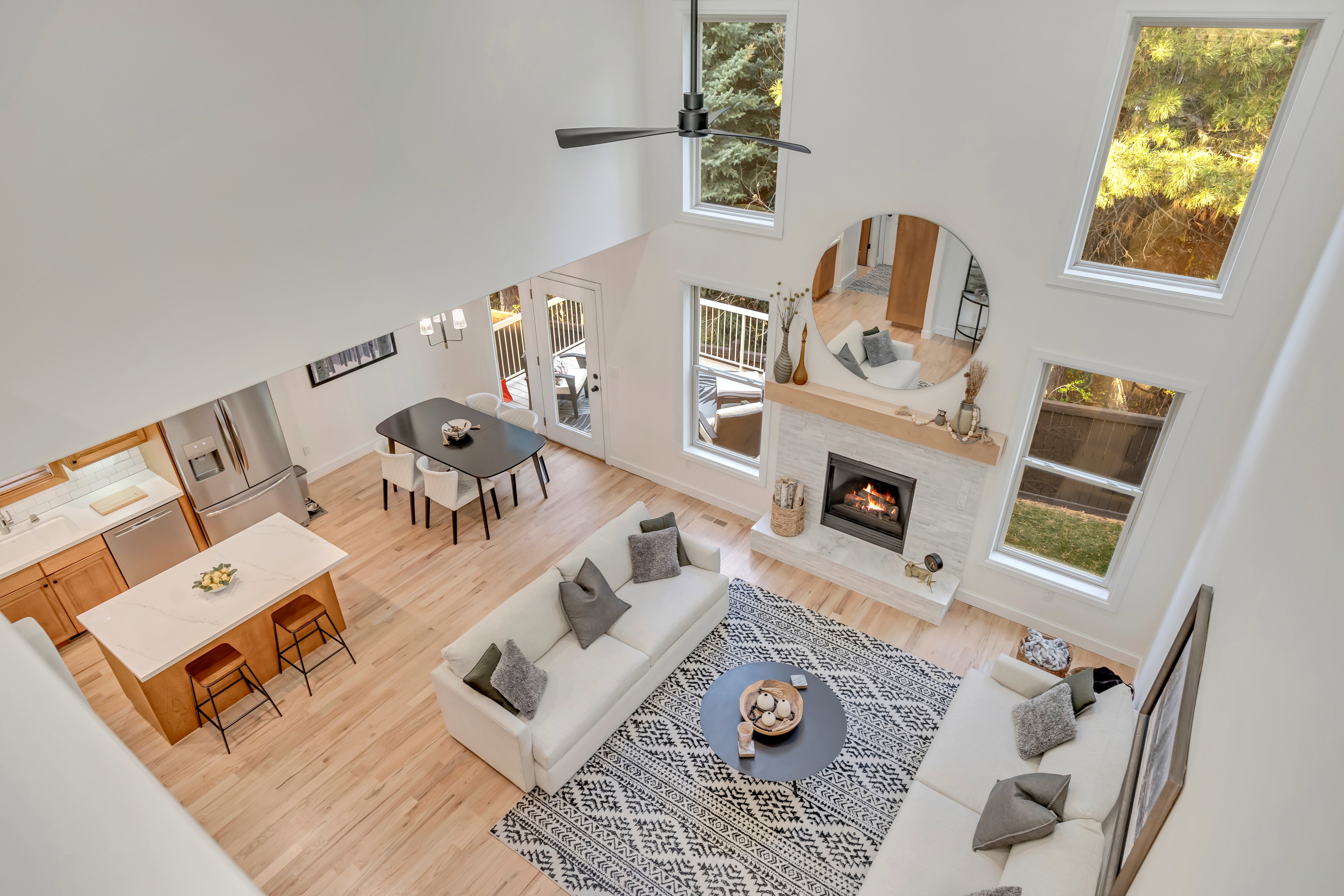Open-concept living space featuring vaulted ceilings, abundant natural light from multiple windows, a modern fireplace with a light-toned tile surround and wood mantle, light wood-finish flooring, and a contemporary kitchen with a central island and stain