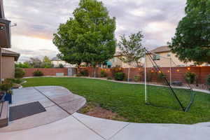 Expansive backyard featuring a manicured lawn, concrete patio, mature trees, a red brick fence, and string lighting