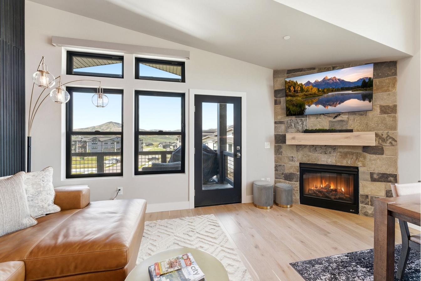 Living area featuring wide plank wood-finish flooring, a stone-clad fireplace with a wood mantel, a glass-paned exterior door, and multiple windows with black frames