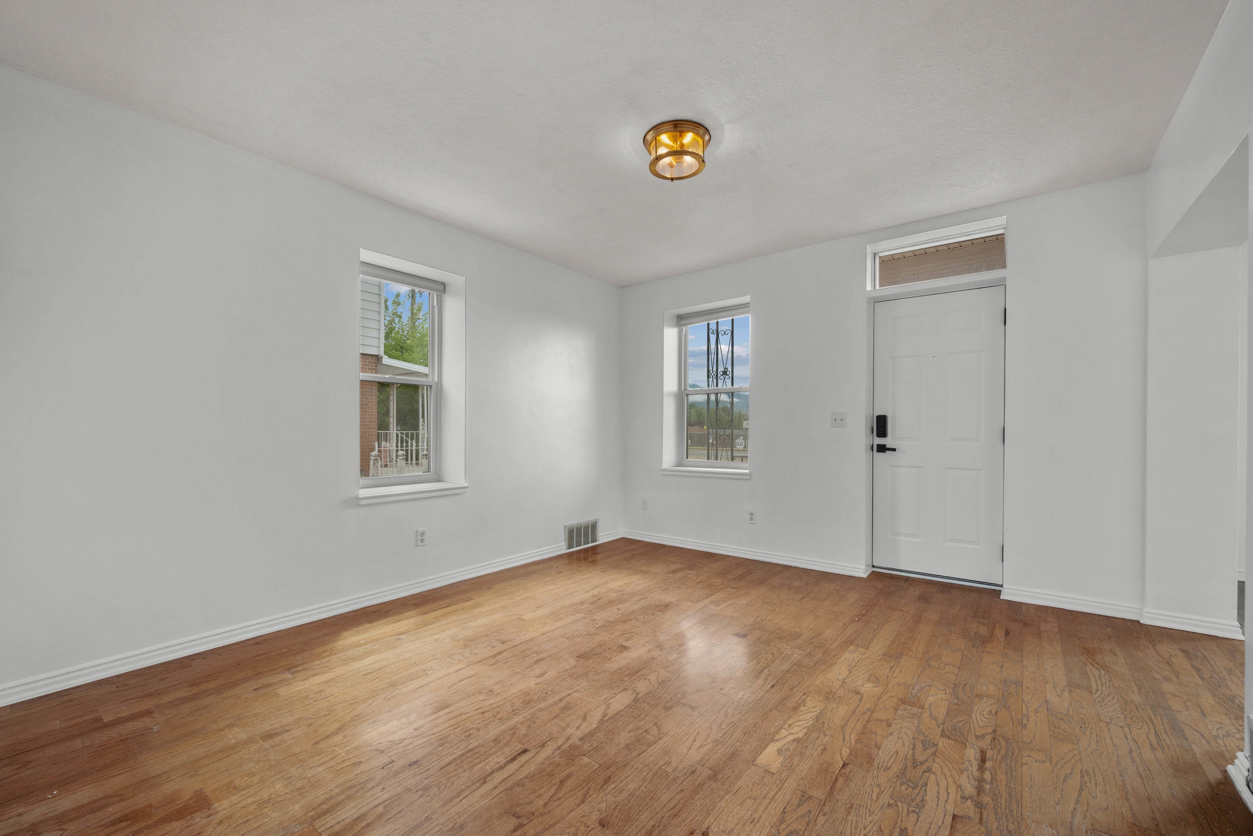 Hardwood flooring throughout a bright room featuring white walls, two windows, and a white entry door with an overhead transom
