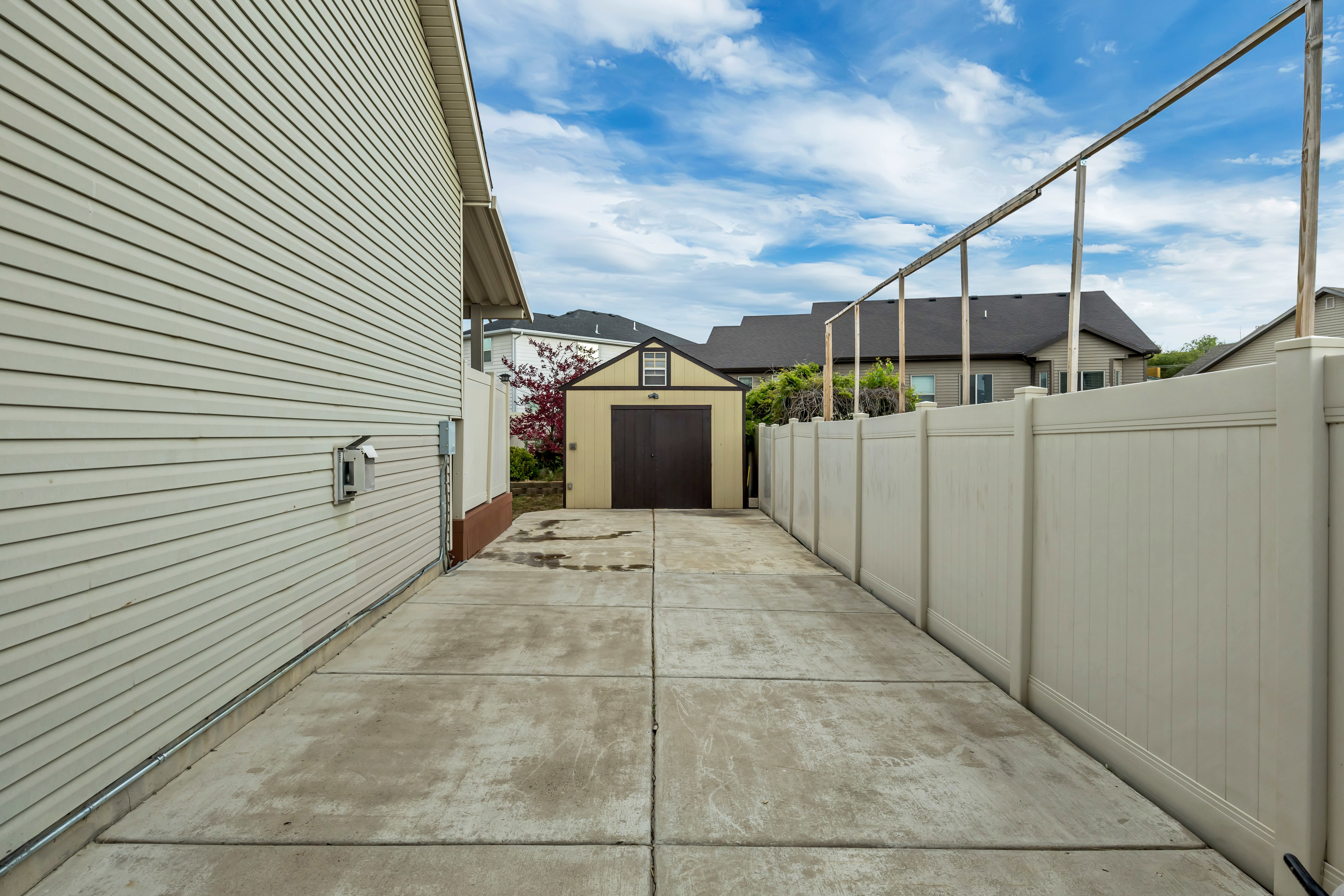 Concrete driveway leading to a detached storage shed with a brown door and white trim, bordered by a light-colored privacy fence