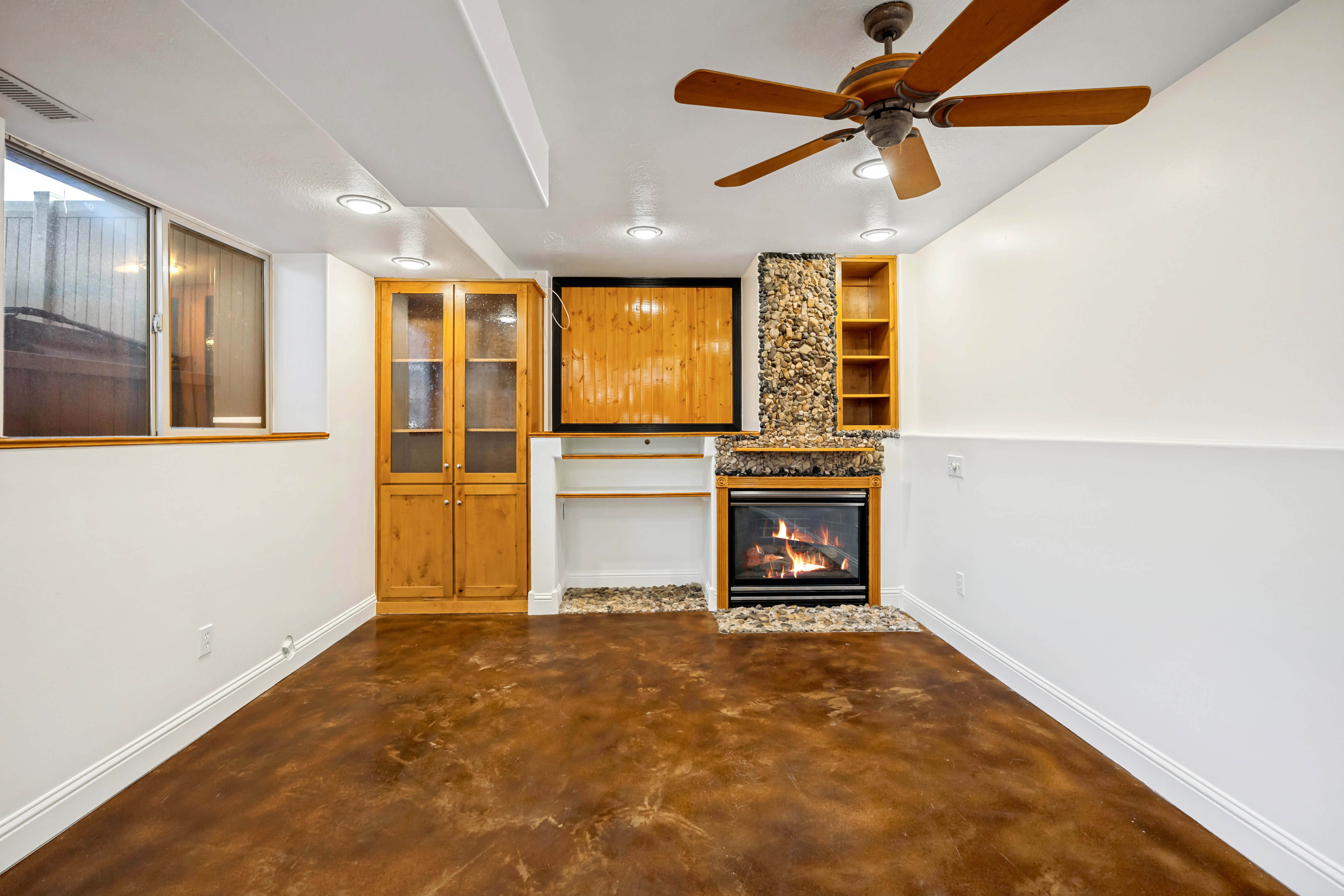 Spacious room featuring concrete flooring, a fireplace with a stone surround, built-in shelving, and a ceiling fan