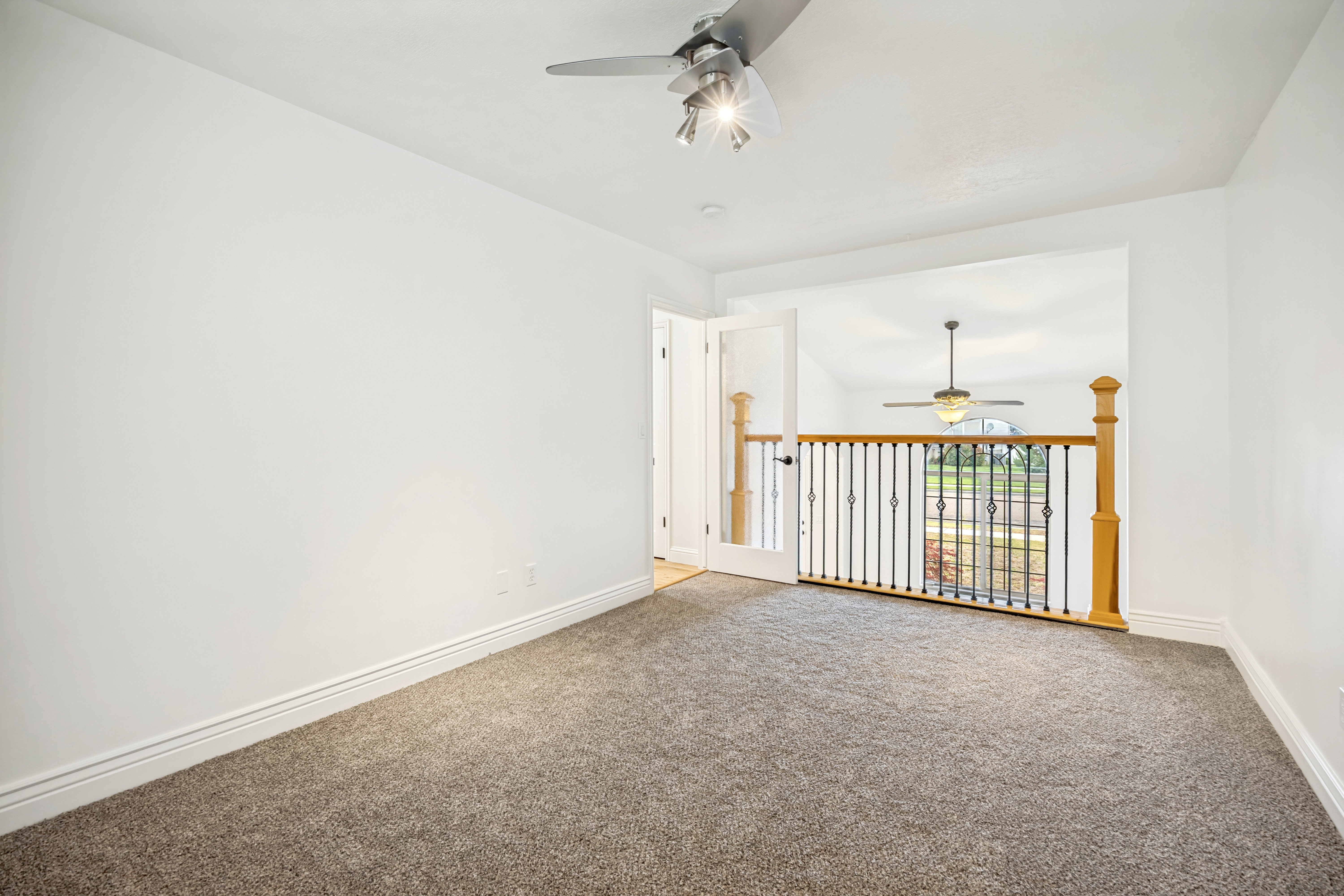 Carpeted room with white walls, a ceiling fan with track lighting, and a doorway opening to a railing with decorative metal spindles