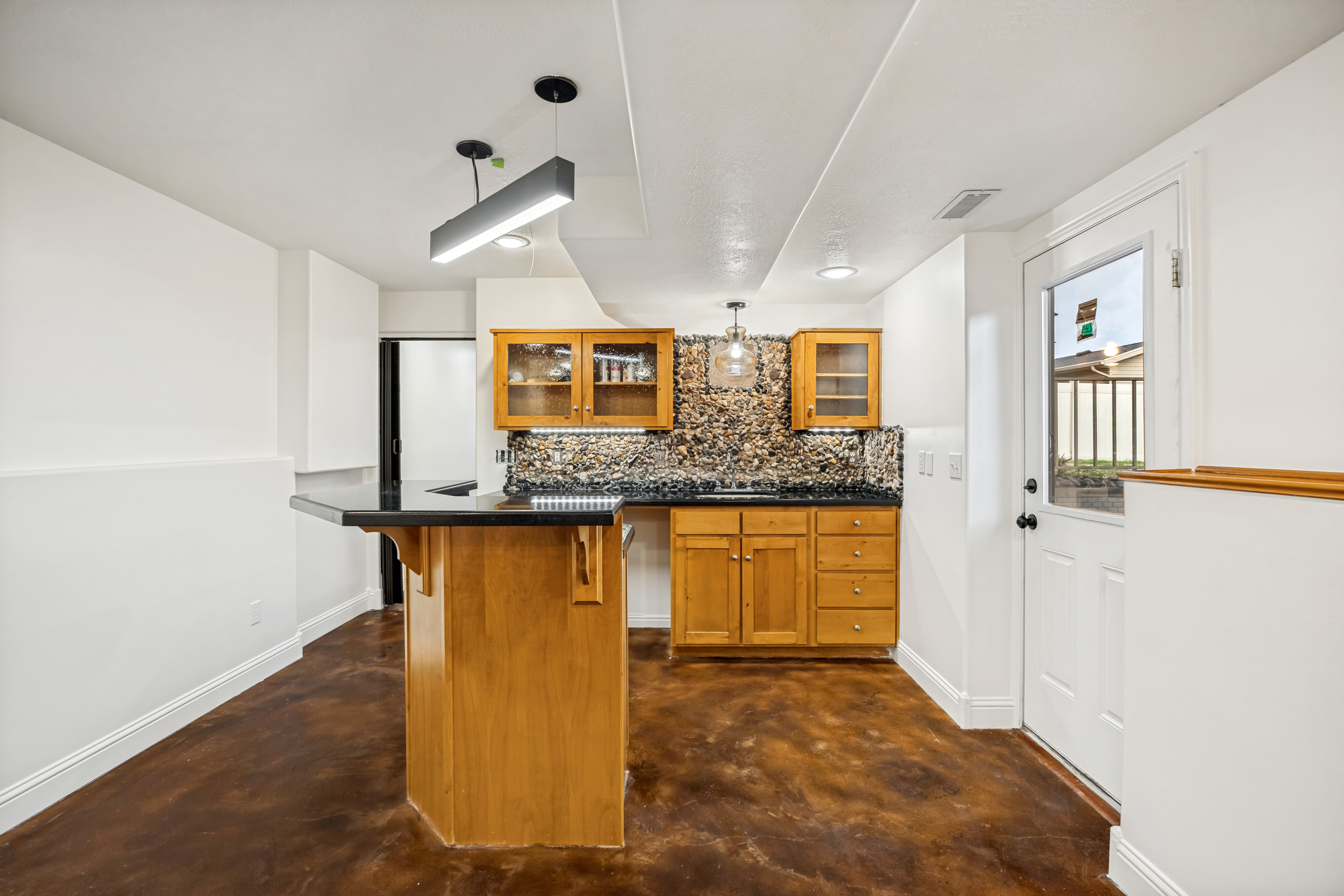 Wood-finish flooring anchors this space featuring a kitchen with a stone tile backsplash, wood cabinetry with glass door accents, and a black countertop
