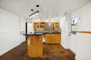 Wood-finish flooring anchors this space featuring a kitchen with a stone tile backsplash, wood cabinetry with glass door accents, and a black countertop