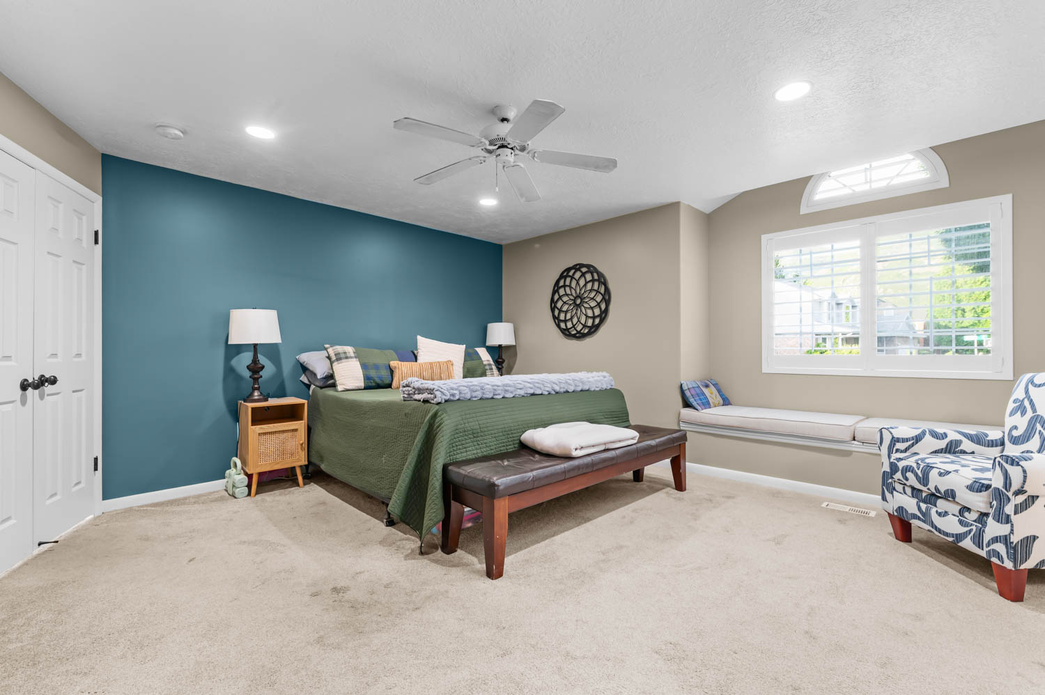 Carpeted bedroom featuring a window seat with cushions, a large window with plantation shutters, an arched transom window, a ceiling fan with integrated lighting, and recessed ceiling lights