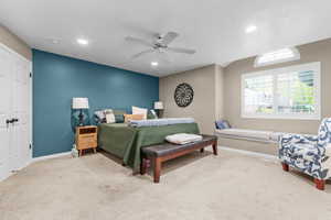 Carpeted bedroom featuring a window seat with cushions, a large window with plantation shutters, an arched transom window, a ceiling fan with integrated lighting, and recessed ceiling lights