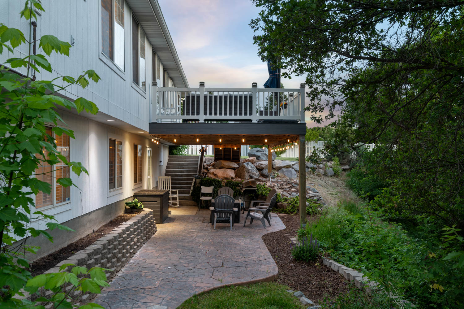 Expansive stamped concrete patio featuring string lighting, terraced landscaping with natural rock elements, and a concrete block retaining wall