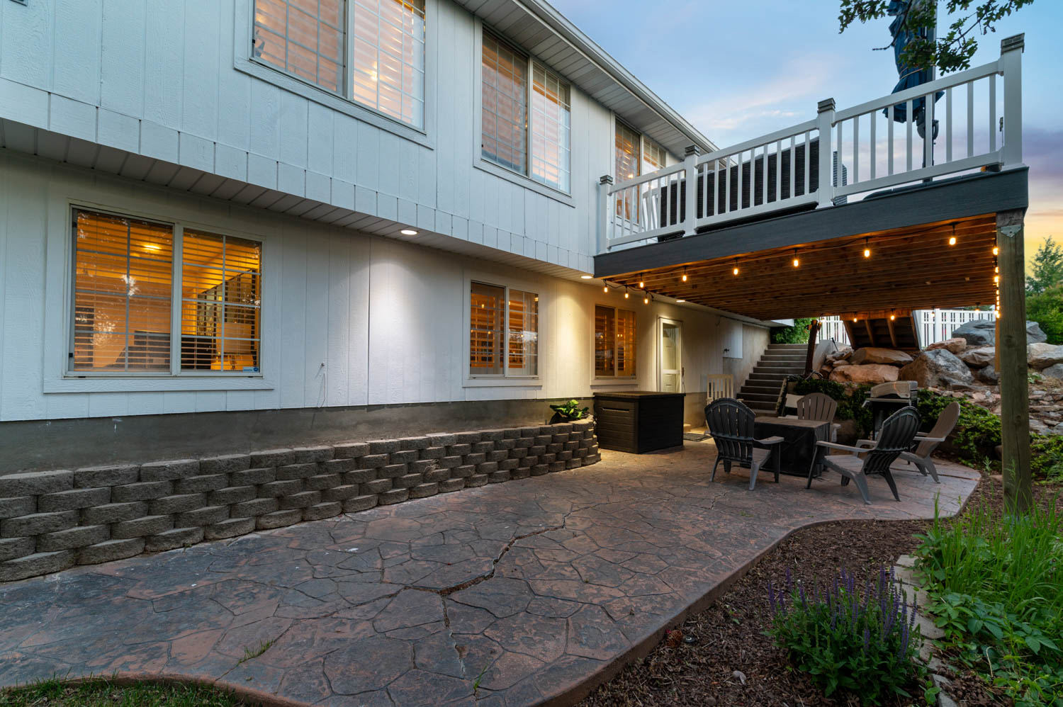 Expansive stamped concrete patio with overhead string lighting, featuring a built-in retaining wall and a staircase ascending to an upper deck