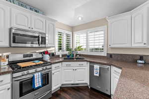 Kitchen featuring white cabinetry, stainless steel appliances, dark stone-finish countertops, wood-finish flooring, and white plantation shutters