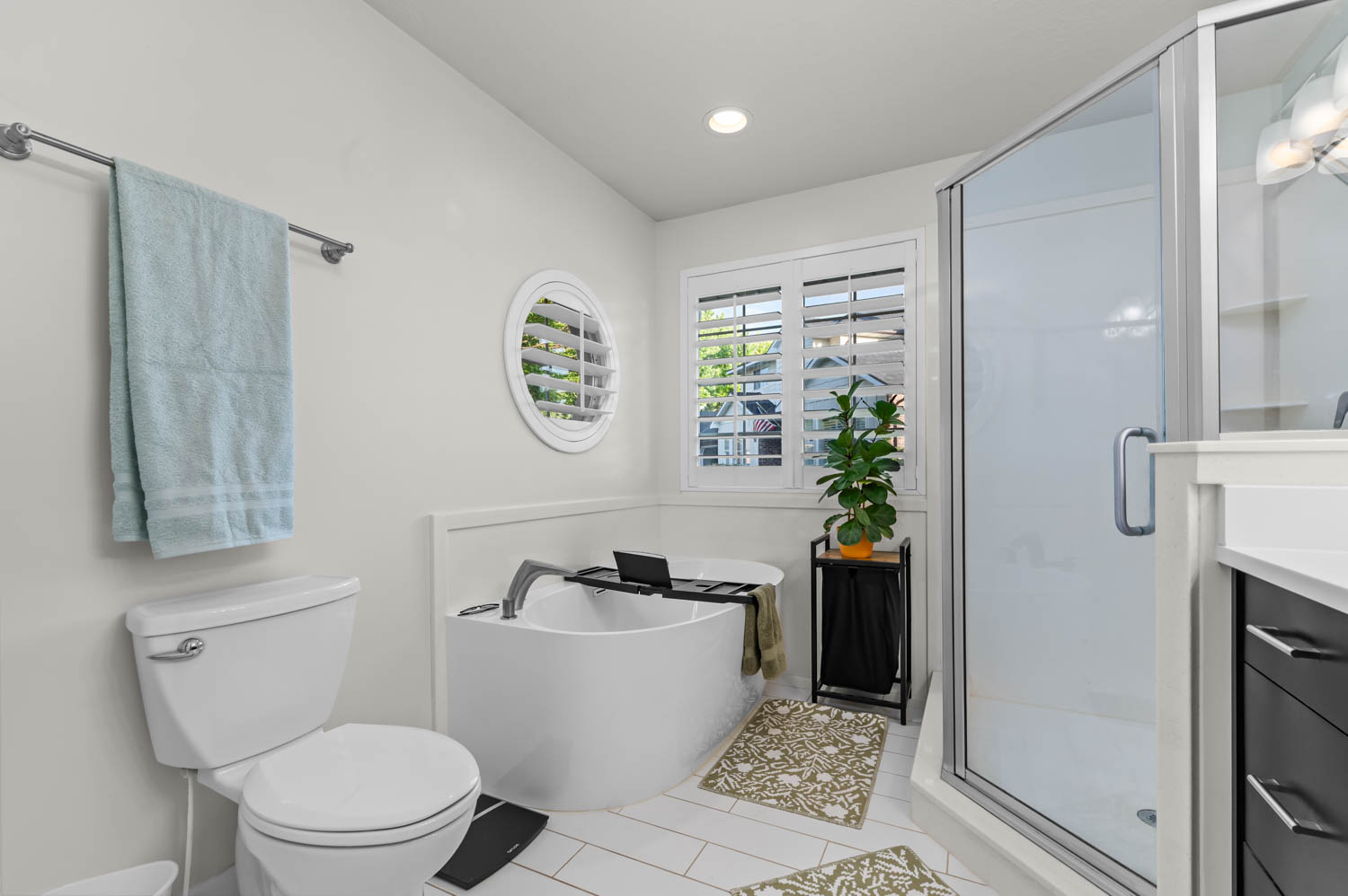Contemporary bathroom featuring a freestanding tub, glass-enclosed shower, rectangular window with plantation shutters, circular accent window, and white tile flooring