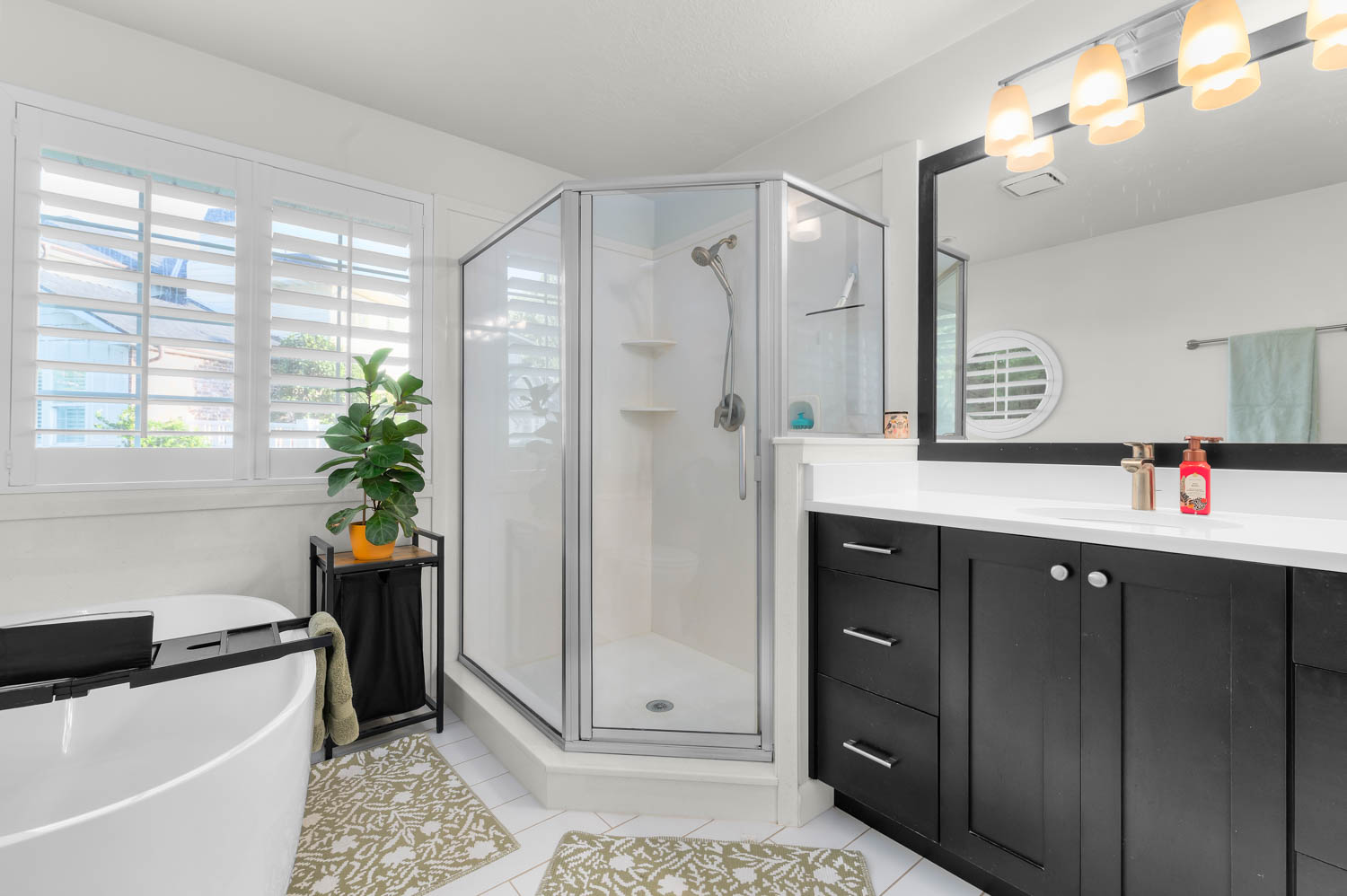 Modern bathroom featuring a standalone corner shower with glass enclosure, a freestanding soaking tub, dual windows with plantation shutters, a dark wood-finish vanity with a white countertop, and a large wall-mounted mirror with an overhead light fixture