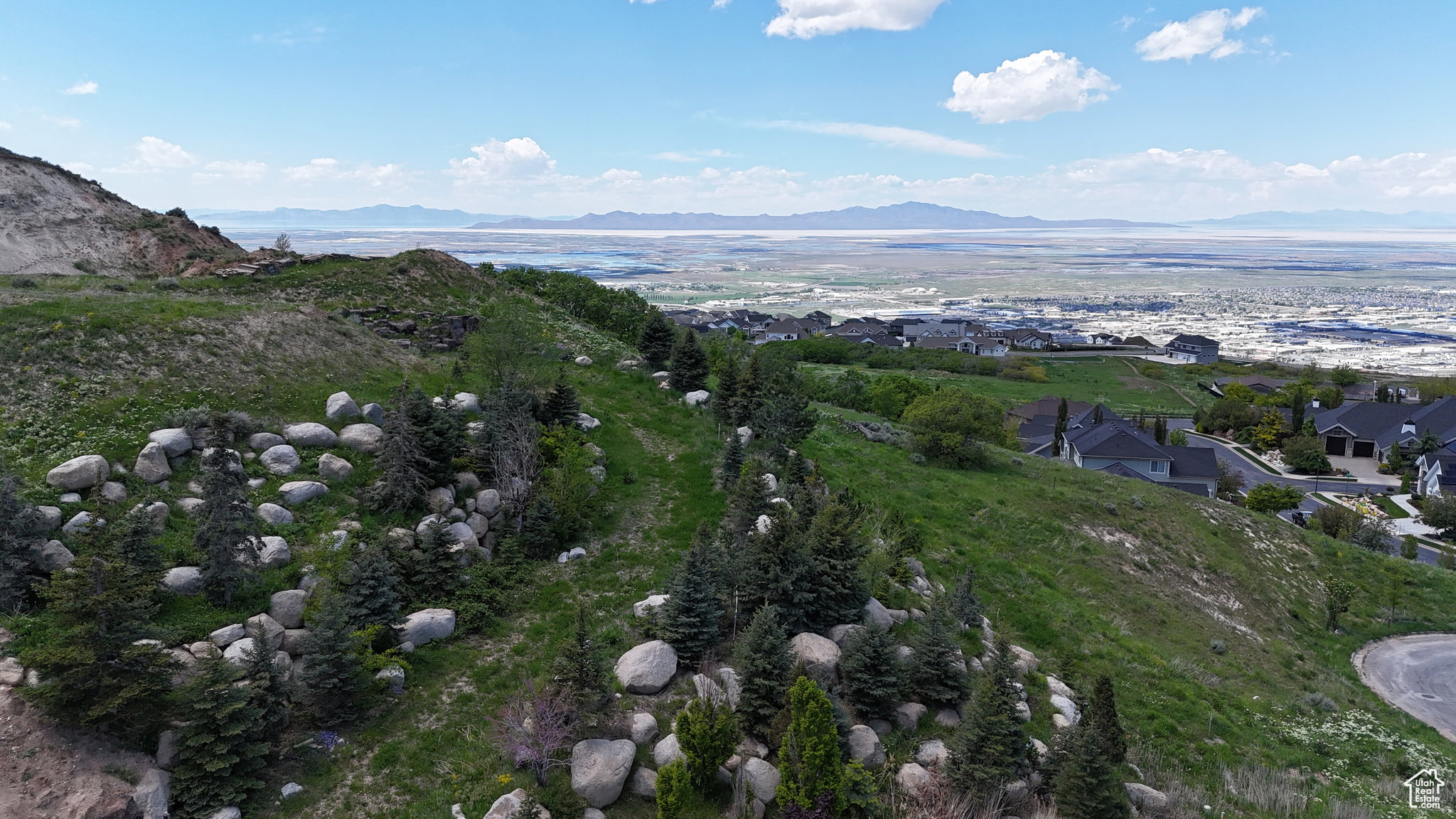 Elevated terrain featuring scattered boulders and evergreen trees with distant residential structures and a large body of water under a clear sky