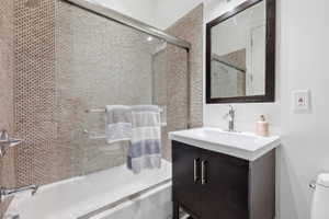 Bathroom featuring a dark wood-finish vanity with an integrated white sink, a framed mirror, and a tiled shower-bath combination with a glass enclosure