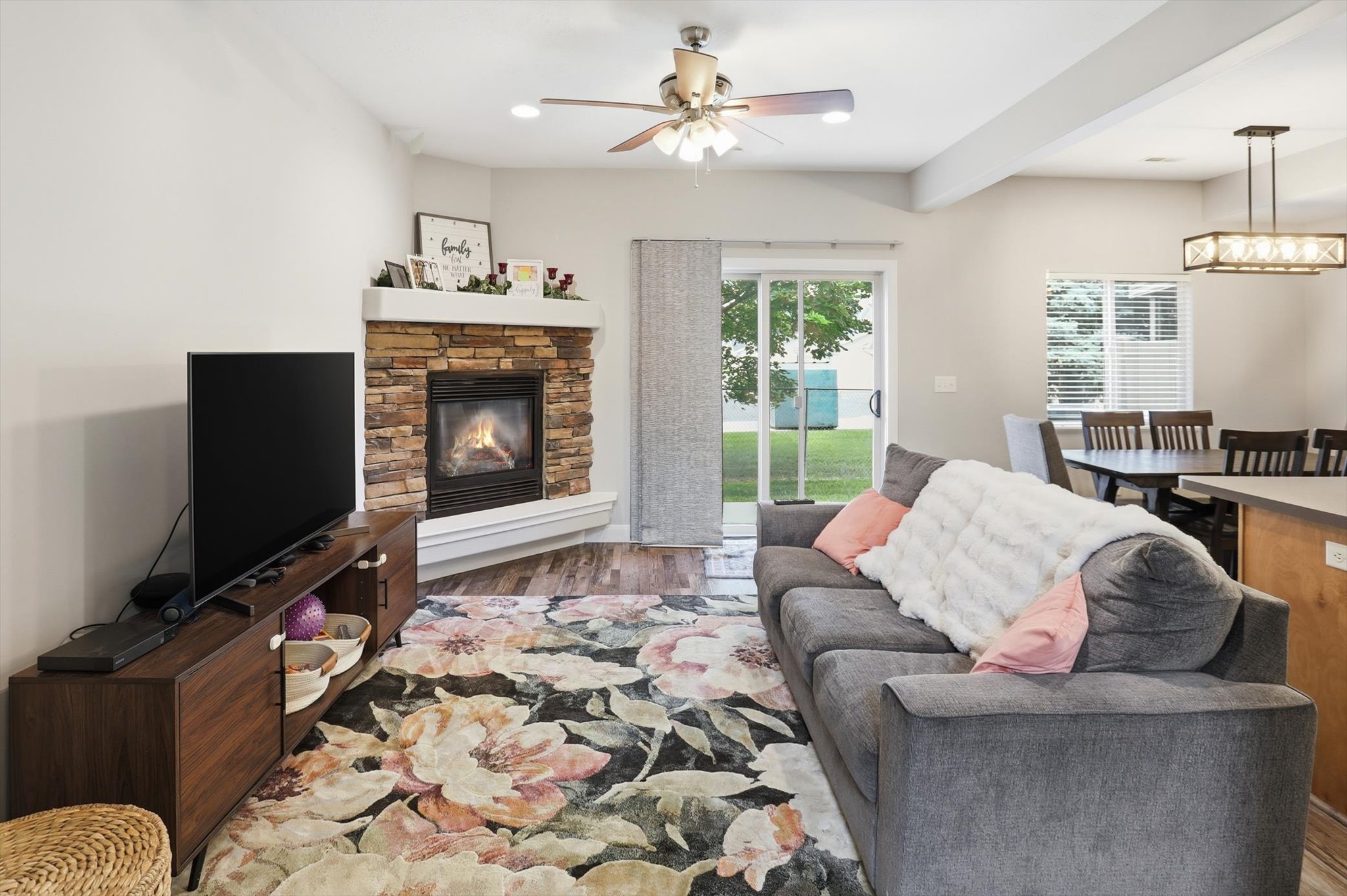 Cozy living space featuring a stone-clad corner fireplace with a white mantel, wood-finish flooring, and a ceiling fan
