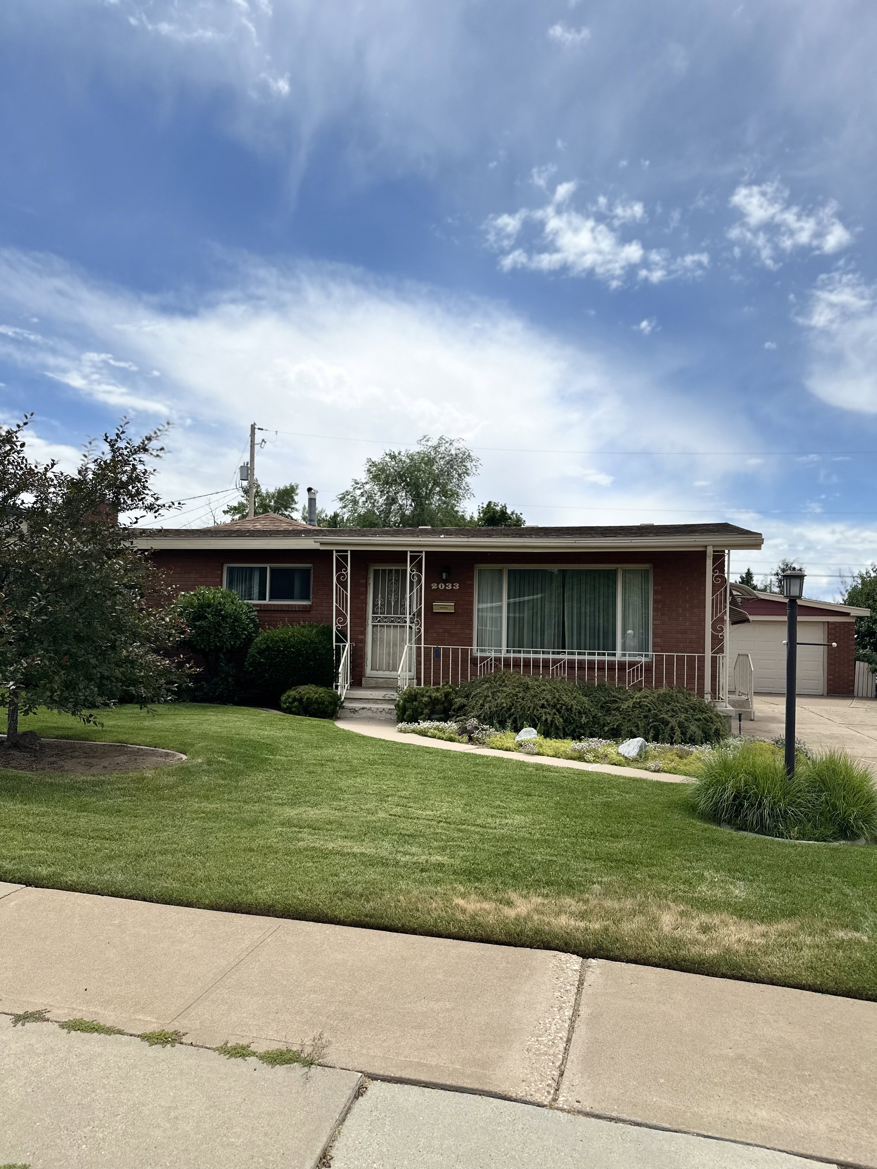 Brick-clad residence featuring a covered front entry with decorative railing, an expansive front lawn, mature landscaping, and a detached two-car garage