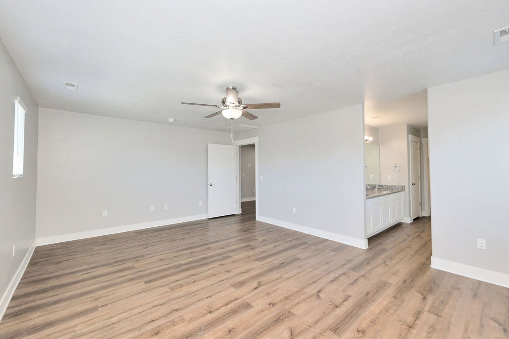 Spacious room featuring wood-finish flooring, light gray walls, a ceiling fan with integrated lighting, and white trim