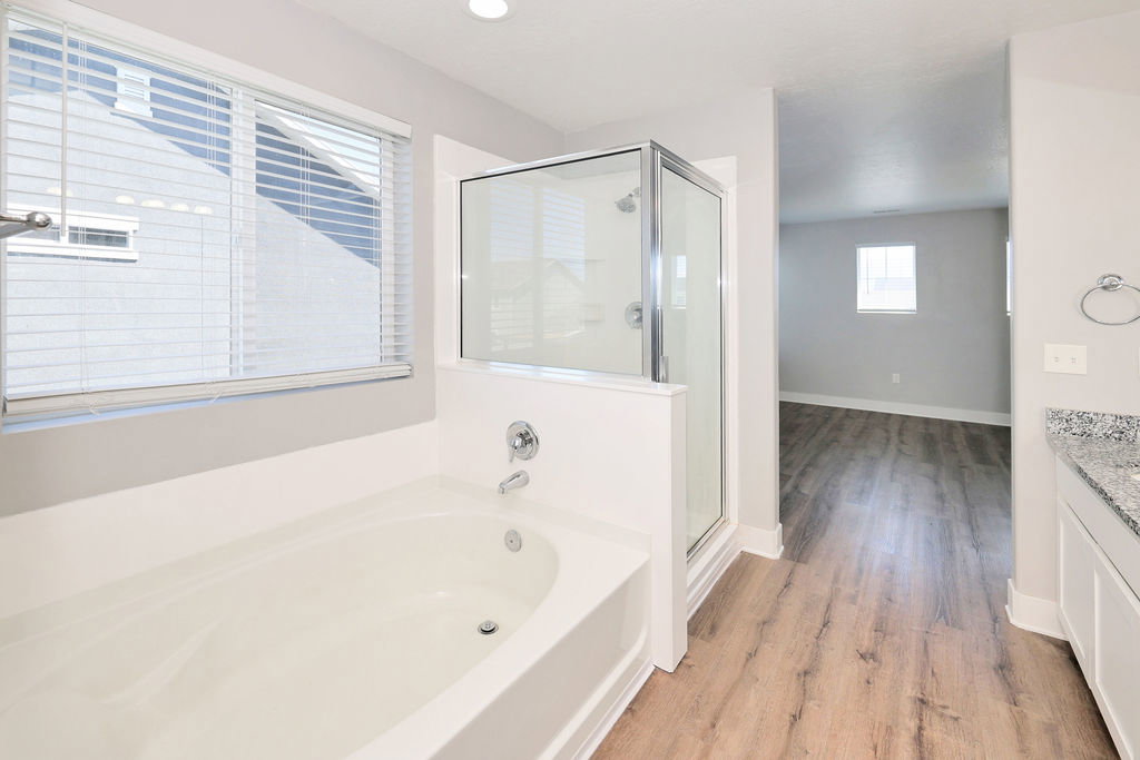 Bathroom featuring a built-in soaking tub, a glass-enclosed shower, wood-finish flooring, and a vanity with a speckled countertop