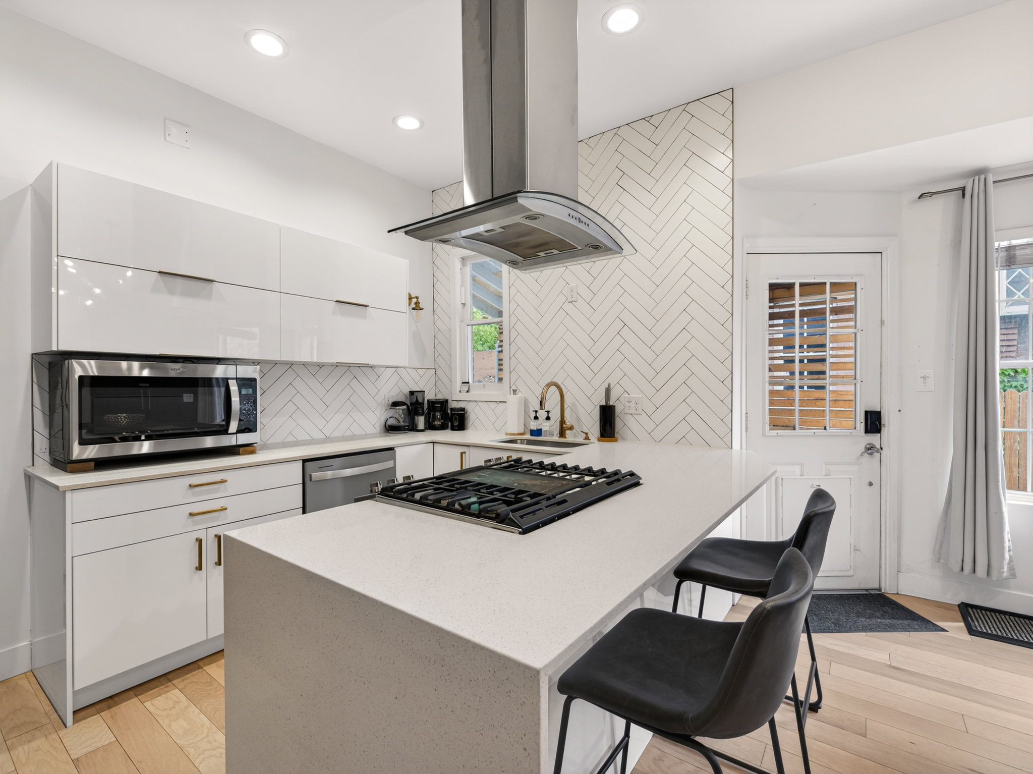 Modern kitchen featuring white high-gloss cabinetry, light wood-finish flooring, a white herringbone tile backsplash, and a large central island with a built-in gas cooktop