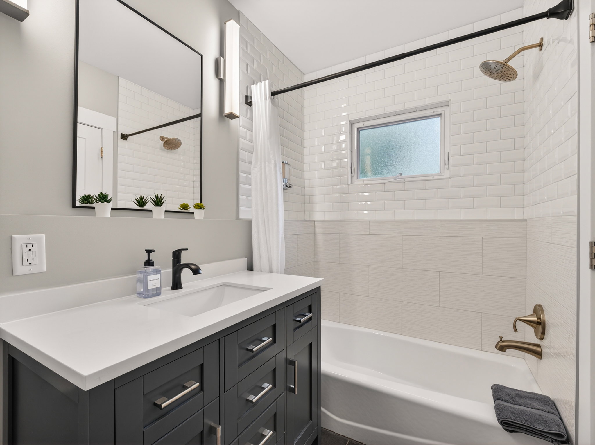Bathroom featuring a dark wood-finish vanity with a white countertop and undermount sink, black-framed mirror with vertical wall sconces, white subway tile shower surround, and a bronze-finish showerhead and tub faucet