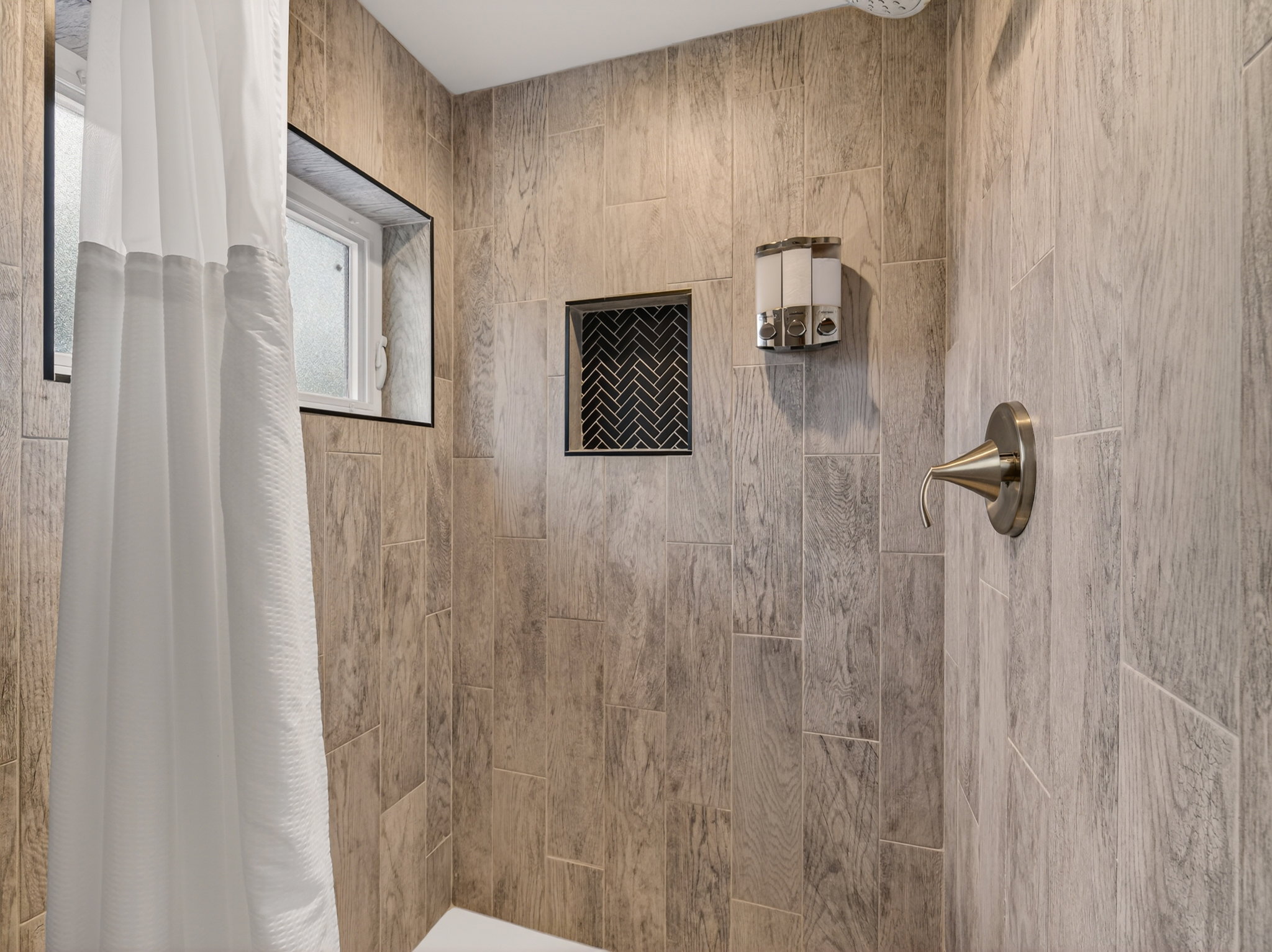 Shower enclosure featuring wood-finish tile, a built-in niche with herringbone tile, an integrated window, a brushed nickel shower fixture, and a white shower base