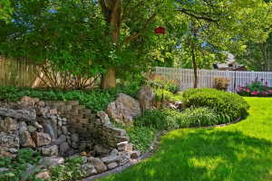 Landscaped backyard featuring a tiered stone waterfall, stone steps, mature trees, vibrant plantings, and a white picket fence